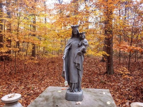 A stone statue of a woman holding a child, surrounded by autumn foliage and fallen leaves in a serene forest setting.
