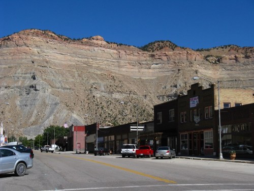 A small town street with shops and parked cars, set against a backdrop of a large, rocky mountain. Clear blue sky above.