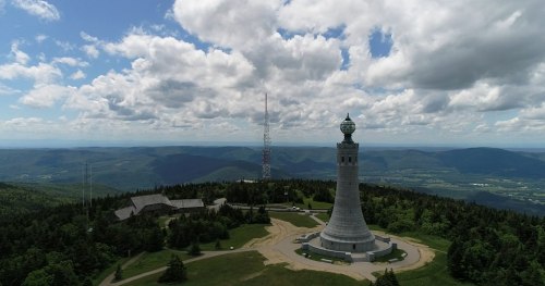 A tall monument with a globe on top, surrounded by trees and mountains under a cloudy sky.