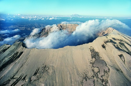Aerial view of a volcanic landscape with clouds surrounding the peaks and valleys, showcasing rugged terrain and blue sky.