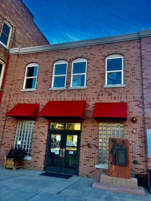 Brick building with red awnings, large windows, and a stone monument in front. Clear blue sky above.