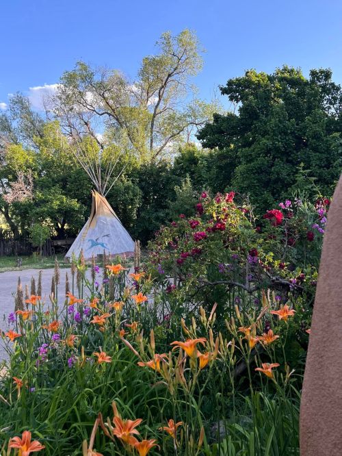 A teepee surrounded by vibrant flowers and lush greenery under a clear blue sky.