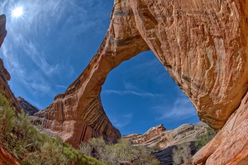 A stunning rock arch framed by blue sky and sunlight, surrounded by layered red rock formations and greenery below.