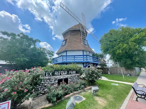A windmill structure at Tex Ritter Park, surrounded by greenery and flowers under a blue sky with fluffy clouds.
