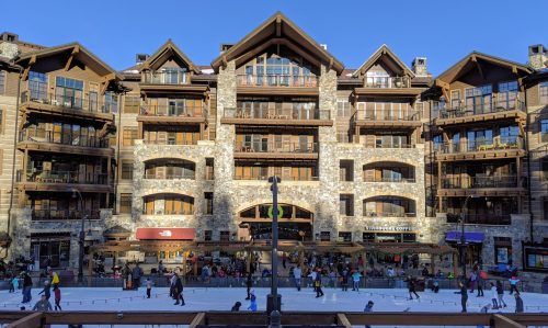 A bustling plaza with people skating and shopping in front of a large, rustic-style building with balconies.