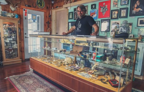 A man stands behind a display case filled with various collectibles in a quirky, decorated shop.