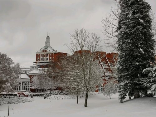 A snowy landscape featuring a historic red brick building and bare trees, with a cloudy sky overhead.