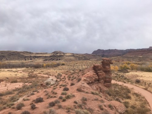 A rocky outcrop stands in a desert landscape with sparse vegetation and distant hills under a cloudy sky.