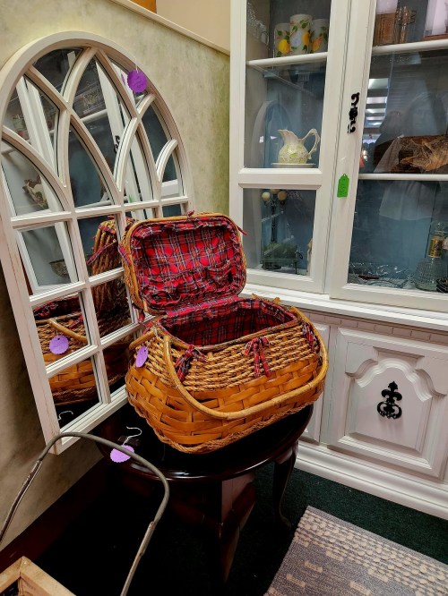 A wicker basket with a plaid lining sits open on a table, reflecting in a decorative mirror nearby.