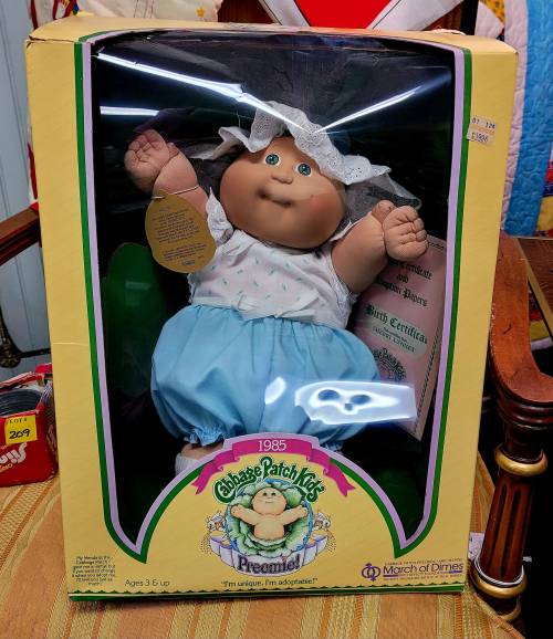 A Cabbage Patch Kids doll in a box, wearing a blue outfit and a white bonnet, labeled "Preemie!" from 1985.