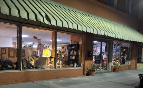 A cozy shop window at night, showcasing various items, a lamp, and decorative plants under a striped awning.