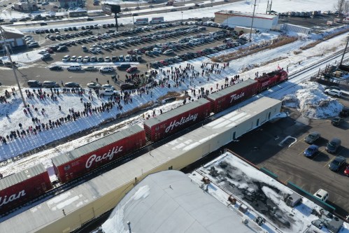 Aerial view of a train with holiday-themed cars passing by a crowd in a snowy area, with parked cars nearby.