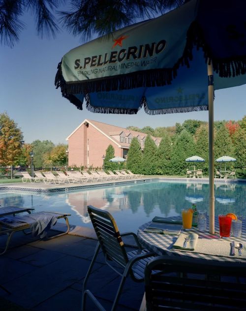 A serene poolside scene with lounge chairs, umbrellas, and a view of a brick building surrounded by trees.