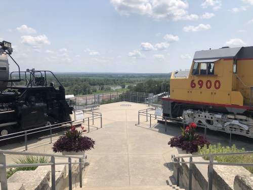 Two vintage trains displayed on a platform with a scenic view of greenery and a river under a blue sky.
