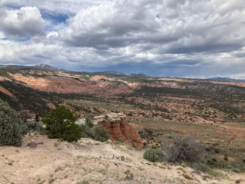 A scenic view of rugged, colorful cliffs and valleys under a cloudy sky, with sparse vegetation in the foreground.