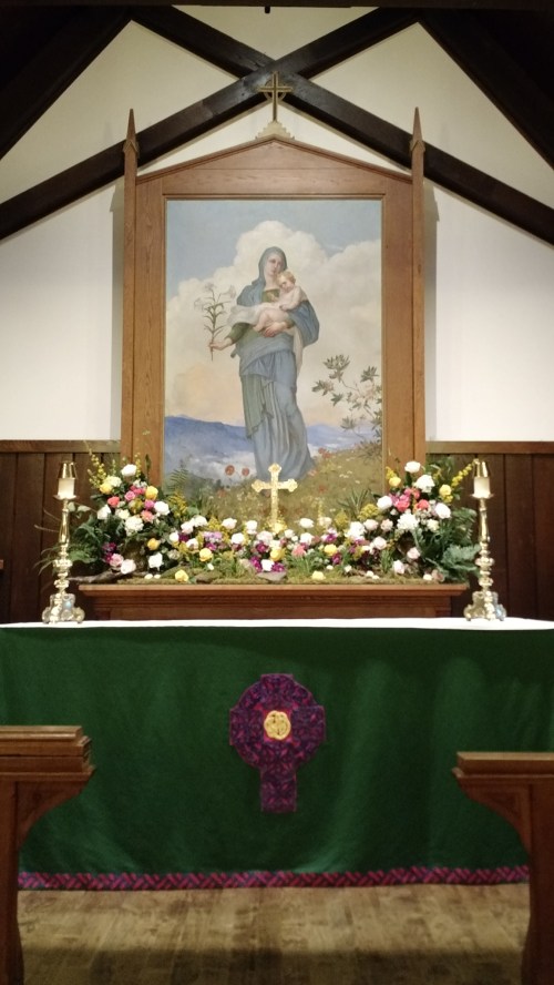 Altar with a floral arrangement, a cross, and a painting of a woman holding a child in a church setting.