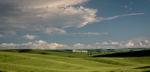 Lush green fields under a blue sky with clouds, featuring distant silos on the horizon.