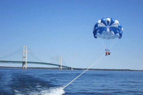 Two people parasailing over water with a bridge in the background under a clear blue sky.