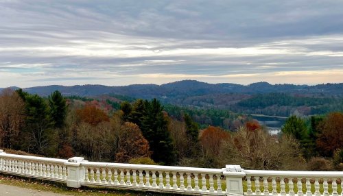 Scenic view of rolling hills and trees with autumn foliage, framed by a white railing under a cloudy sky.