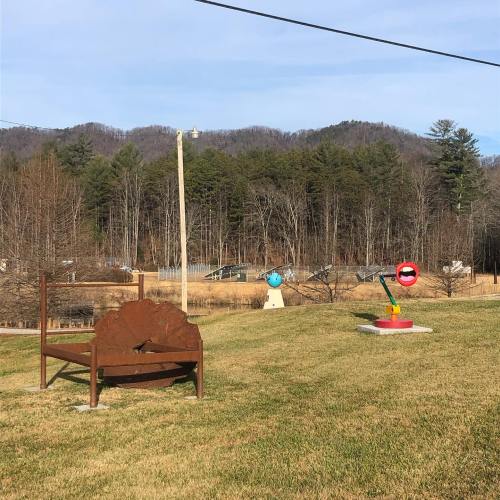 A grassy area with a wooden bench, colorful sculptures, and a backdrop of trees and mountains under a clear sky.