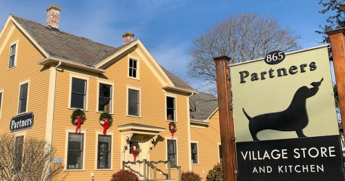 A yellow house with festive decorations and a sign for "Partners Village Store and Kitchen" in front.