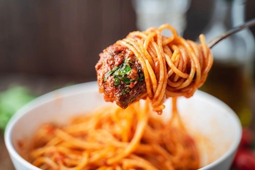 A fork holding spaghetti with meatballs and marinara sauce, above a bowl of pasta.