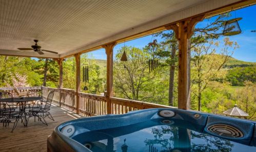 A spacious porch with a hot tub, dining table, and scenic views of green hills and trees under a clear blue sky.