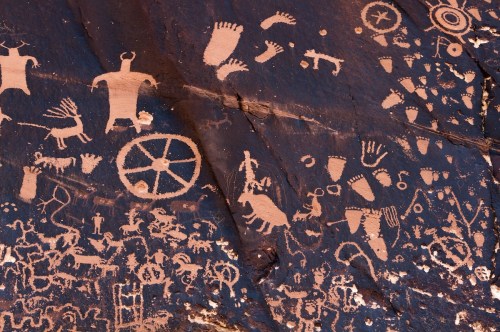 Petroglyphs on a rock surface, featuring human figures, animal shapes, and various symbols in a desert landscape.