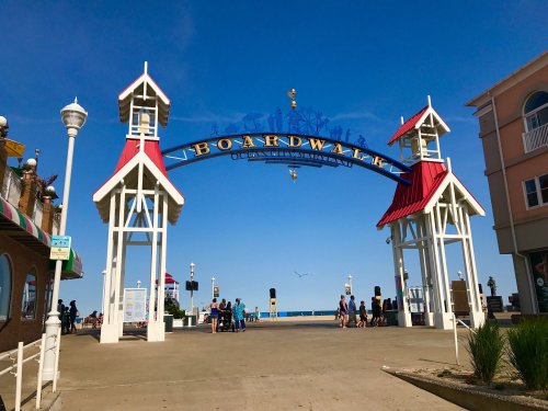 A sunny boardwalk entrance with a colorful arch, leading to the beach and ocean, bustling with visitors.