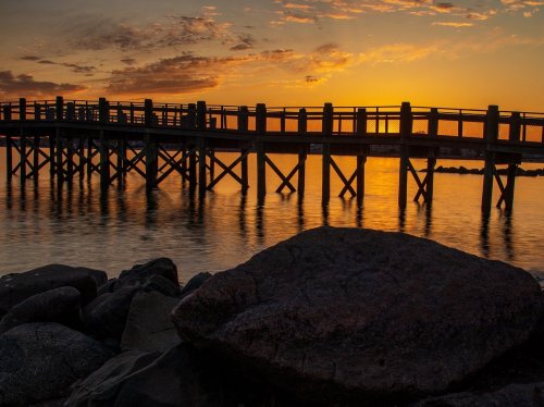 A wooden pier extends over calm water at sunset, with rocks in the foreground and a colorful sky.
