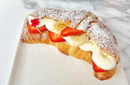 A flaky croissant filled with cream, strawberries, and banana, dusted with powdered sugar on a white plate.