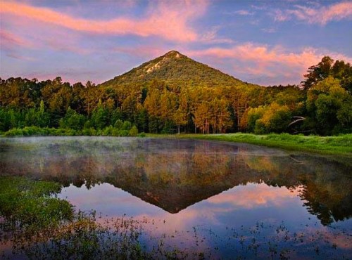 A serene lake reflects a mountain at sunset, surrounded by lush trees and a colorful sky.