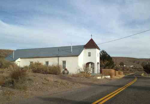 A small white church with a gray roof, set against a desert landscape along a winding road.