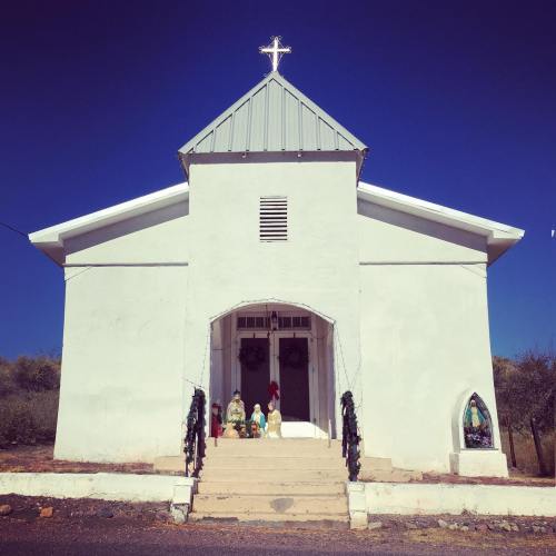 A white church with a cross on top, featuring a nativity scene on the steps and a statue of Mary to the right.