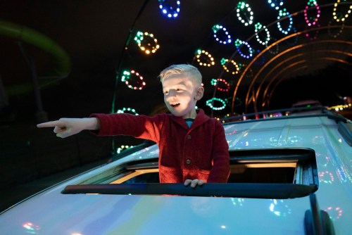 A young boy in a red sweater joyfully points out of a car sunroof, surrounded by colorful lights at night.