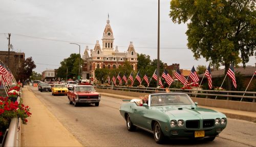 A vintage car drives past a historic building adorned with flags, with other cars on a street lined with flowers.