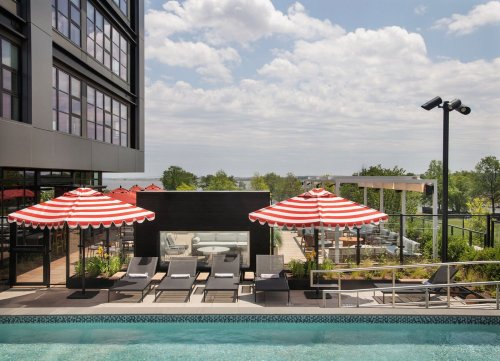 A pool area with striped umbrellas, lounge chairs, and a modern building in the background under a partly cloudy sky.