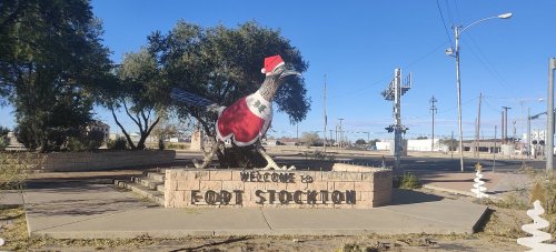 A large bird statue wearing a Santa hat, with a "Welcome to Fort Stockton" sign in a sunny outdoor setting.