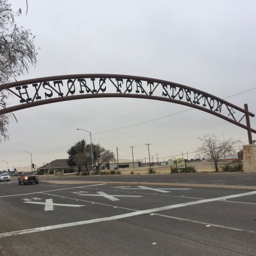 Archway sign reading "Historic Fort Stockton" over a road, with a cloudy sky and sparse landscape in the background.