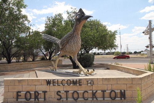 A large statue of a roadrunner stands at the entrance to Fort Stockton, with trees and a road in the background.