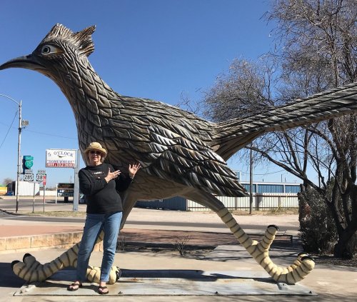 A person poses next to a large, whimsical statue of a roadrunner in a sunny outdoor setting.