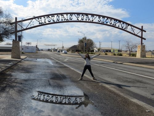 A person stands joyfully under the "Historic Fort Stockton" archway, with a puddle reflecting the sign.