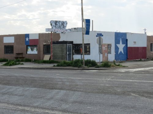 A weathered building with Texas flag murals on its exterior, located at a street corner. Grass and weeds are visible.