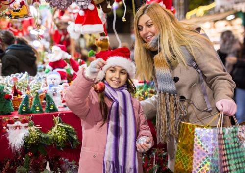 A mother and daughter smile while shopping at a festive market, holding ornaments and surrounded by holiday decorations.