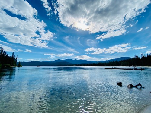 A serene lake surrounded by mountains under a bright blue sky with fluffy clouds, reflecting on the water's surface.