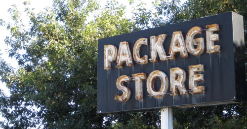 Sign for a package store with weathered letters, surrounded by green trees.