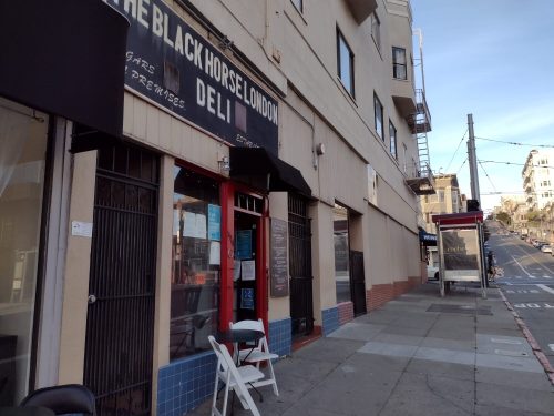 Exterior of The Black Horse London Deli, featuring a red entrance and sidewalk seating, on a sloped street.