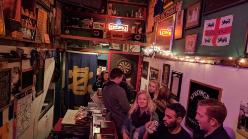 A lively bar scene with patrons chatting, a dartboard, and colorful decorations on the walls.