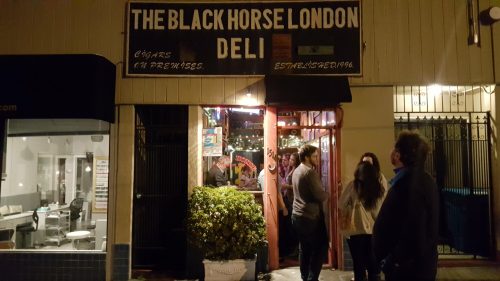 A night scene of The Black Horse London Deli, with people gathered outside the entrance.