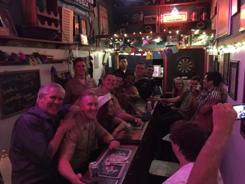A lively bar scene with a group of people smiling and posing for a photo, drinks in hand, and colorful decorations.